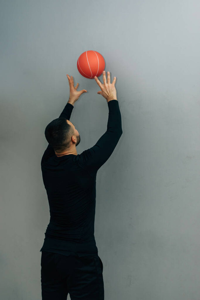 Joven atleta masculino agachado haciendo ejercicios de bolas de pared en el gimnasio sobre fondo gris. - Foto, Imagen