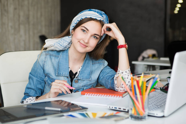 Porträt einer jungen schönen lächelnden, lässig lächelnden Designerin am Schreibtisch mit Instrumenten, Farbmustern und Laptop - Foto, Bild