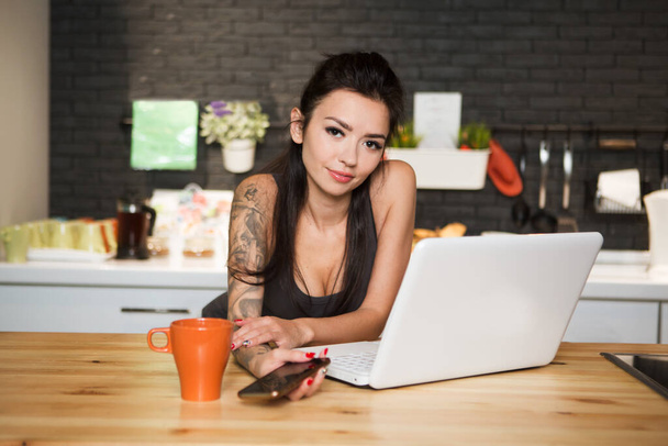 Beautiful young woman with coffee cup and laptop in the kitchen at hom. home office. - Foto, immagini
