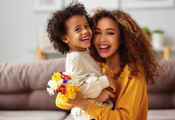 Возбужденный мальчик маленький сын поздравляет свою маму счастливой смешанной расовой женщиной с Днем матери и дарит ей цветочный букет, они обнимаются и смеются, сидя дома на диване. Концепция семейного отдыха - Фото, изображение
