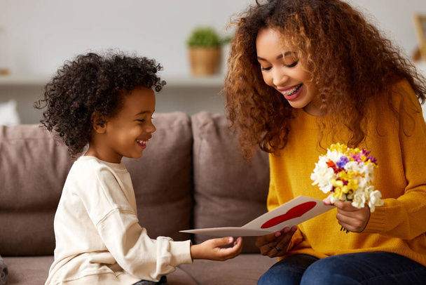 With love. Cute excited little boy son giving his mom handmade greeting postcard and flower bouquet, happy mother embracing with with child while sitting on sofa at home. Mothers day celebration concept - 写真・画像
