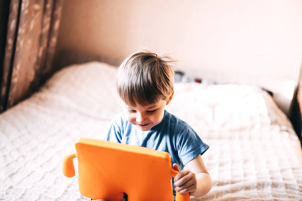 Mały chłopiec siedzi na łóżku i trzyma tabliczkę w rękach. Uczenie się dzieci na odległość. Nowoczesna edukacja domowa dla dzieci. - Zdjęcie, obraz