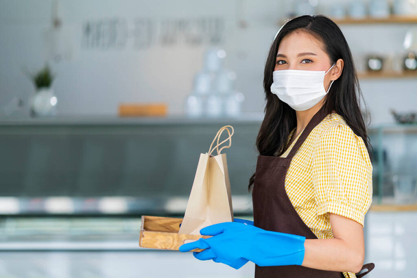 Atrakcyjne azjatyckie kobieta kawiarnia pracownik nosi maskę twarzy i rękawice dając na wynos żywności torba do customer.kelnerka gospodarstwa zamówienie na wynos stojąc w kawiarni drzwi wejściowe nowy normalny styl życia - Zdjęcie, obraz
