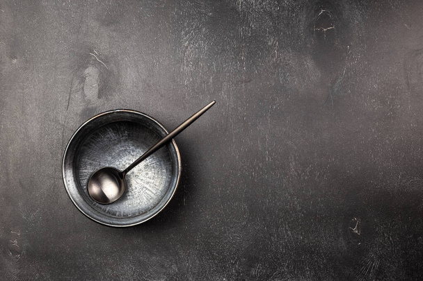 Κενό μαύρο πήλινο πιάτο μπολ και μαύρο μεταλλικό κουτάλι που σε ξύλινο τραπέζι. Μονοχρωματικά χρώματα - Φωτογραφία, εικόνα