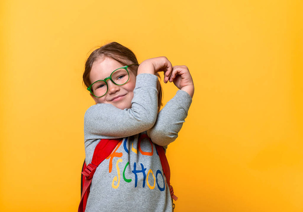 Happy smiling kid girl making heart sign. Advertising gesture, charity, donation, like and education. Kid with backpack and glasses over yellow background. Back to school. - Fotó, kép