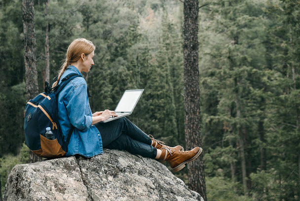 Young woman with backpack sitting on ground at forest and working on laptop . Workation concept. Freelancer work - Foto, Bild