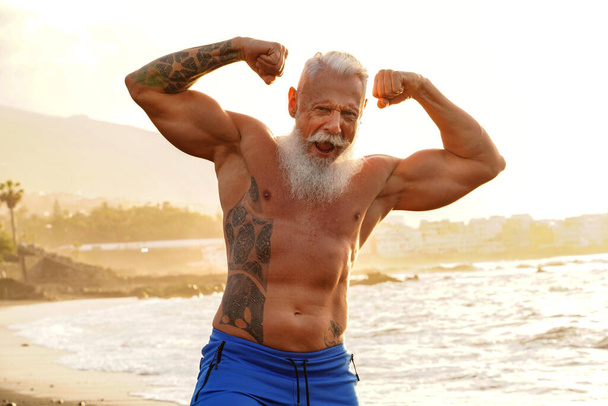 Happy fit senior man showing his muscle on the beach after outdoor workout, during sunset time. In a healthy body healthy mind. Age is just a number. Elderly people lifestyle and real human emotions concept. - 写真・画像