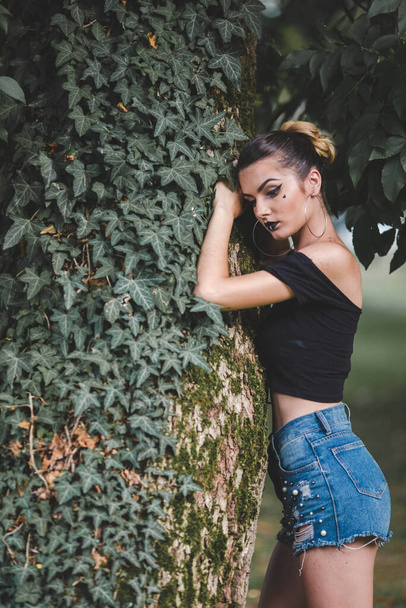 A young attractive Caucasian female in jeans shorts leaning on the tree and posing - Fotoğraf, Görsel