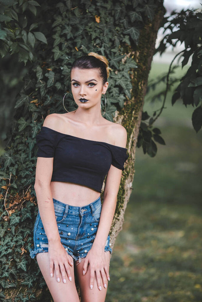 A young attractive Caucasian female in jeans shorts leaning on the tree and posing - Foto, Imagem