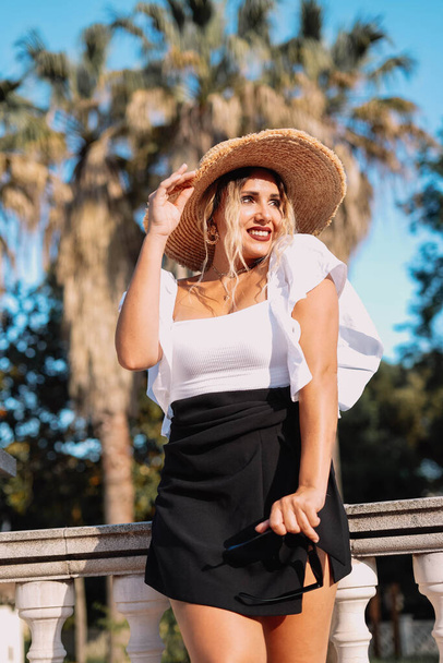 Egy gyönyörű nő képe, aki fekete szoknyában, fehér ingben és nyári kalapban pózol. - Fotó, kép