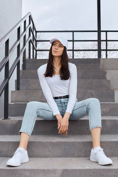 Beautiful young adult woman wearing jeans, white shirt and baseball cap sitting on stairs outdoors and looking away, having serious facial expression, dreams or thinks about important things. - Photo, image