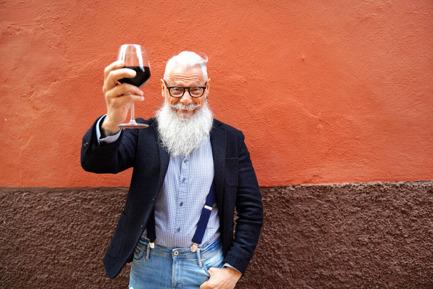 Handsome senior man with white beard and mustache toasting with red wine, looking at camera with big smile. Elegant men celebrating success - 写真・画像