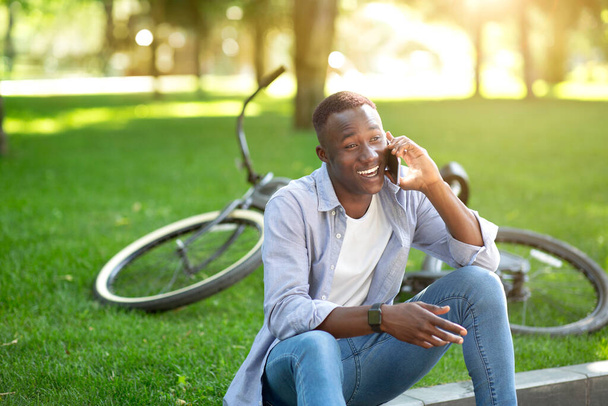 Attractive black man with modern bike speaking on cellphone at green park, space for text - Foto, imagen