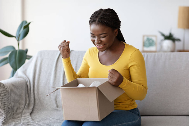 Glückliche schwarze Frau packt Paket zu Hause aus und reagiert emotional auf erfolgreiches Einkaufen - Foto, Bild