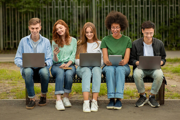 Diversos estudiantes adolescentes sentados con diferentes dispositivos electrónicos en el banco en el parque - Foto, imagen
