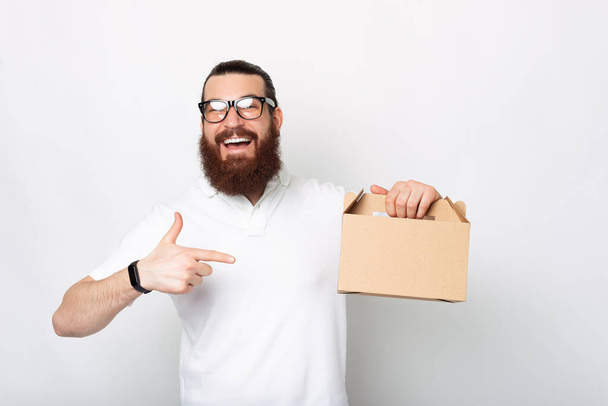 Opgewonden man met een baard draagt een bril wijst naar een lunchbox over een witte achtergrond. - Foto, afbeelding