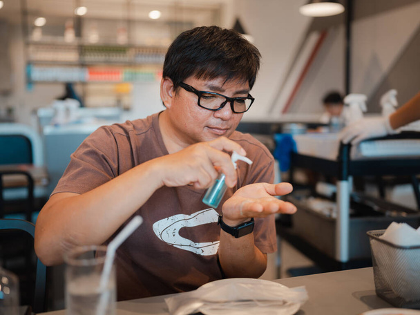 Asian man with eyeglasses using alcohol gel from small bottle to clean hands and protect corona virus or COVID-19 infection before eating in restaurant  - Photo, Image