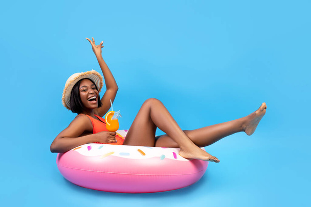 Positive black woman sitting on inflatable ring, holding tropical cocktail on blue background, full length portrait - Fotoğraf, Görsel