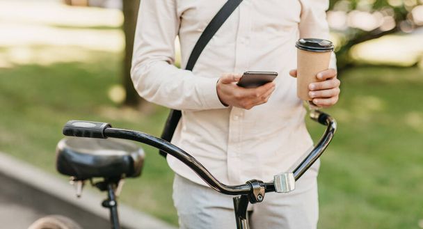 Mladý muž hipster s jízdním kolem drží odnést kávu a psaní zprávy na smartphone v dopoledních hodinách - Fotografie, Obrázek