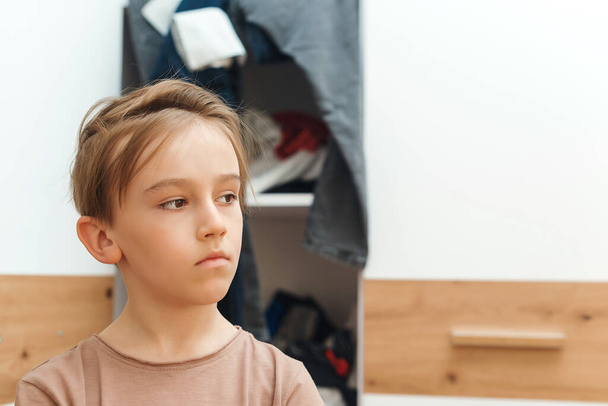 Boy standing near messy clothes on shelf. Preteen boy with dirty clothing in his room. Messy home kid's room. Untidy clutter clothing closet. Boy thinking what to do with messy. - Photo, Image