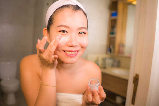 portrait of young happy and beautiful Asian Chinese woman applying facial anti wrinkle cream and serum at bathroom mirror in skin care and beauty treatment concept - Foto, Bild