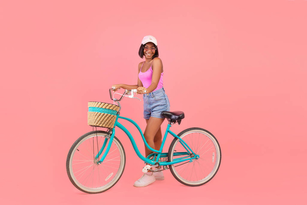 Longitud completa de feliz dama negra en traje de verano de pie con bicicleta clásica sobre fondo de estudio rosa - Foto, Imagen