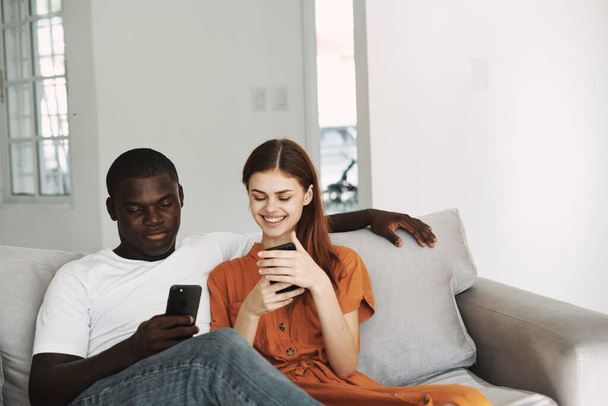 donna in un prendisole e un uomo africano con i telefoni cellulari sono seduti sul divano a chiacchierare interni della famiglia - Foto, immagini