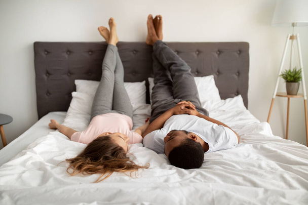 Тысячелетняя межрасовая влюбленная пара, лежащая дома на кровати, поднимающая ноги, разговаривающая друг с другом - Фото, изображение