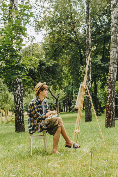 a young guy in a straw hat sits in the park in front of an easel and paints a picture with oil paints. Artistic work in nature. Plein air. - 写真・画像