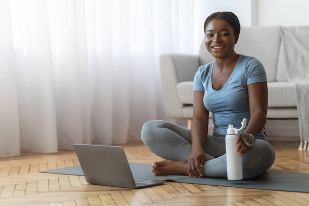 Sorridente donna nera in abbigliamento sportivo seduto con il computer portatile pronto per l'allenamento a casa - Foto, immagini