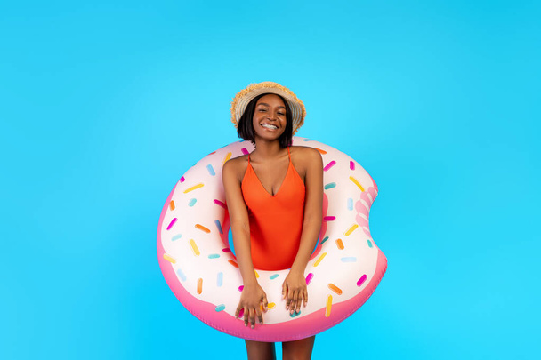 Portrait of joyful millennial black lady in swimsuit posing with inflatable ring on blue studio background - Foto, imagen