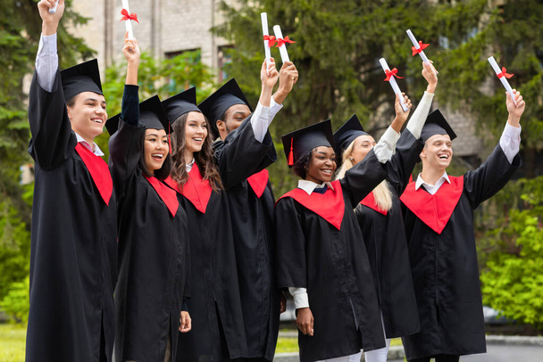 Mezuniyet kostümlü duygusal çok ırklı öğrenciler diplomaları yükseltiyor. - Fotoğraf, Görsel
