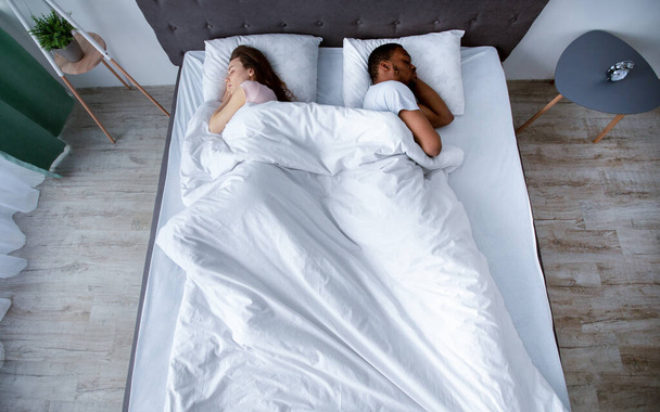 Міленіумна багаторасова пара відвертається один від одного, спить в ліжку вдома, вид зверху
 - Фото, зображення