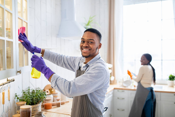 Attractive African American guy cleaning kitchen cabinet with detergent, his wife helping him on background, copy space - Photo, Image