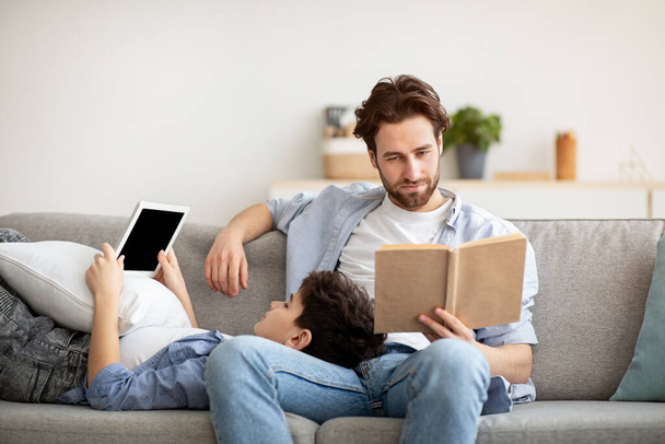 Andere interesses. Jonge vader leest papieren boek terwijl zijn tiener zoon met behulp van digitale tablet, rusten samen op de bank - Foto, afbeelding