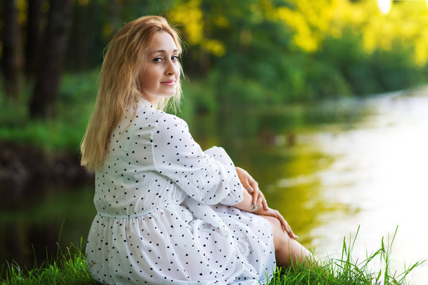 Beautiful young blonde woman in a white dress on the shore of a lake or river on a sunny day. Enjoy your life - Foto, afbeelding