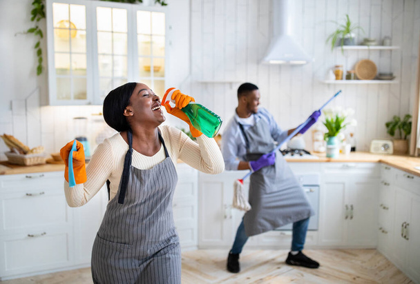 Senhora negra feliz usando detergente como microfone, seu namorado tocando esfregona como guitarra na cozinha, espaço de cópia - Foto, Imagem