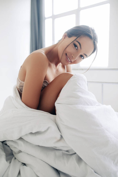 Ελκυστική σέξι γυναίκα κάθεται στο κρεβάτι τυλιγμένη σε κουβέρτα και χαμογελά. - Φωτογραφία, εικόνα