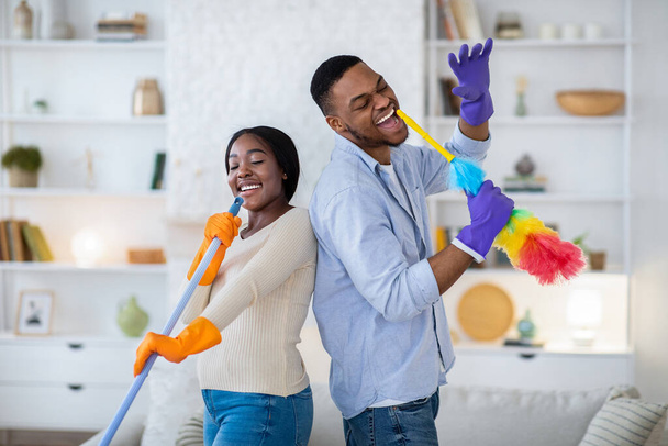 Alegre hombre negro y su novia cantando durante la limpieza de la casa, usando fregona y plumero como micrófonos - Foto, imagen