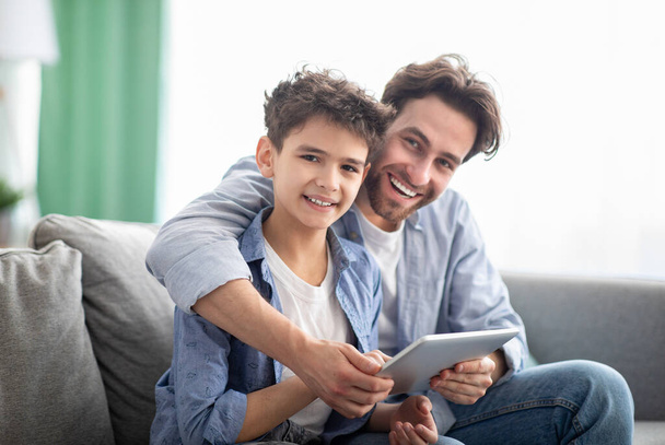 Familie tijd. Gelukkig vader en zoon met behulp van digitale tablet, surfen op internet online thuis, zitten samen op comfortabele bank - Foto, afbeelding