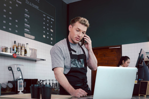 Молодой мужчина-бариста в фартуке, разговаривающий на мобильном телефоне и ноутбуке, стоя за стойкой кафе, обслуживающий персонал кафе. - Фото, изображение