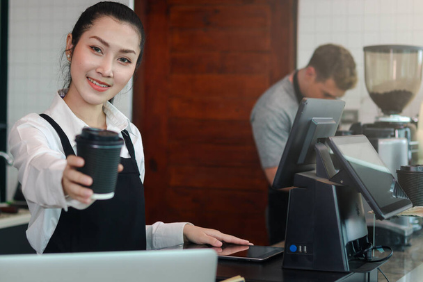 Young beautiful Asian female barista wearing apron, giving paper disposable coffee cups to camera while standing behind coffee shop counter bar, smiling waitress staff working at cafe. - Photo, Image