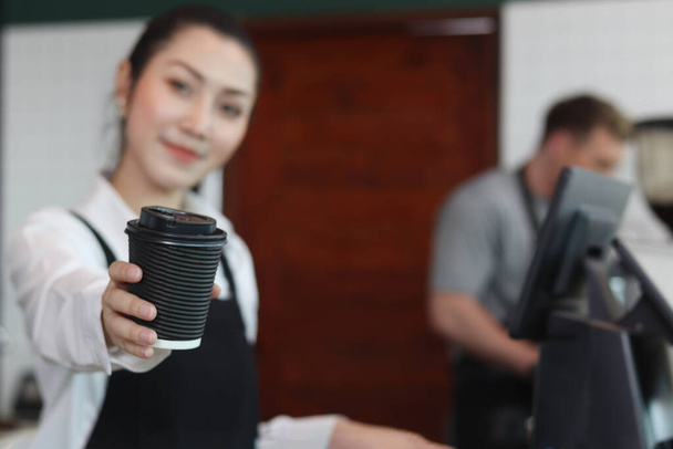Young beautiful Asian female barista wearing apron, giving paper disposable coffee cups to camera while standing behind coffee shop counter bar, smiling waitress staff working at cafe. - Foto, Bild
