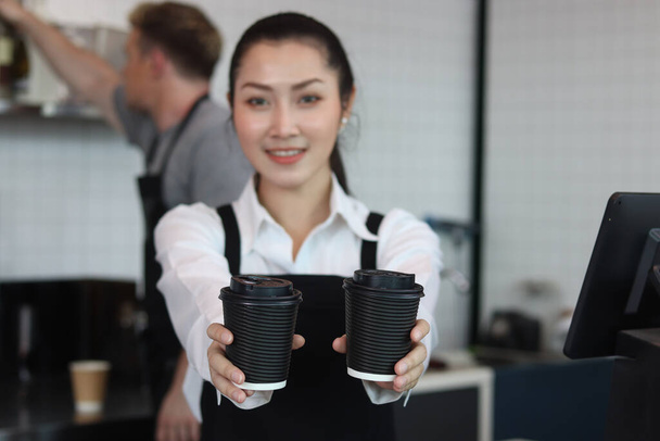 Młoda piękna azjatycka baristka nosząca fartuch, dająca papierowe kubki jednorazowej kawy do kamery stojąc za barem kawiarni, uśmiechnięta obsługa kelnerki pracująca w kawiarni. - Zdjęcie, obraz