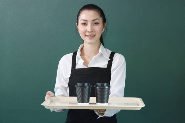 エプロンを身に着けている若い美しいアジアの女性バリスタの肖像、緑の背景に紙使い捨てコーヒーカップのドーディングトレイ、カフェのコンセプトで働くかなりウエイトレスのスタッフを笑顔 - 写真・画像