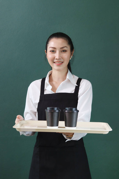 Портрет молодої азіатки бариста у фартусі, ляльковий піднос з паперовими одноразовими чашками кави на зеленому фоні, усміхаючись офіціантці, яка працює в кафе концепт - Фото, зображення