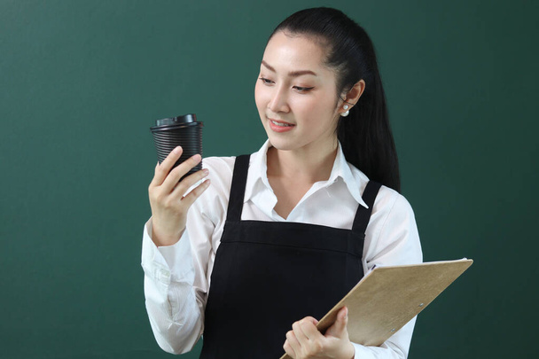 Portret młodej pięknej azjatyckiej baristki ubranej w fartuch, trzymającej menu i papierowy kubek jednorazowej kawy na zielonym tle, uśmiechnięta obsługa kelnerki pracująca przy koncepcji kawiarni. - Zdjęcie, obraz