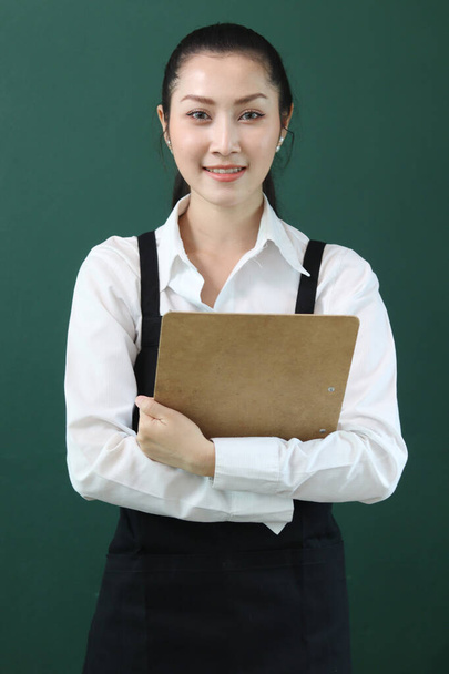 Portrét mladé krásné asijské barista nosit zástěru, držení menu a papírové jednorázové šálek kávy na zeleném pozadí, usmívající se hezká servírka zaměstnanci pracující v kavárně koncept. - Fotografie, Obrázek