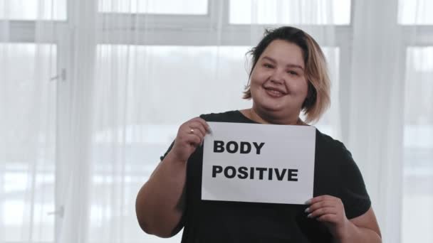 Koncepcja pozytywności ciała - pulchna uśmiechnięta kobieta trzyma znak z napisem BODY POSITIVE - patrząc w aparat - Materiał filmowy, wideo