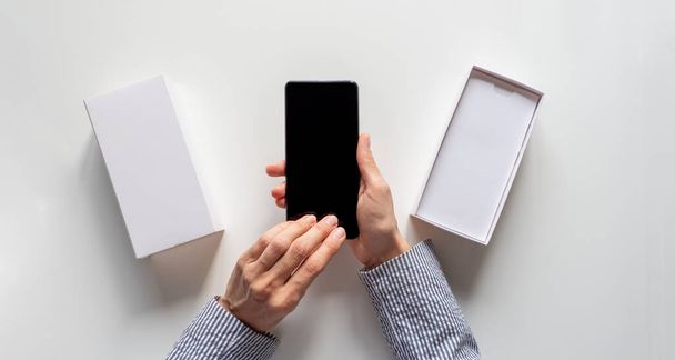 Unpacking a new smartphone on a white table. Female hands holding modern smartphone with blank screen. Online shopping concept. Top view. - Foto, afbeelding